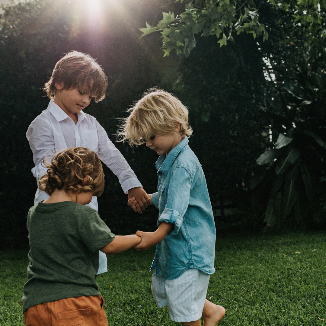 Three boys holding hands in a circle outdoors on the grass. 